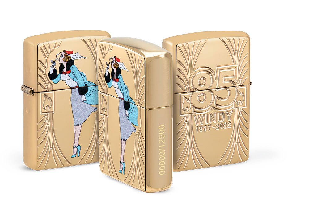 Ein goldfarbenes Zippo-Feuerzeug zeigt eine stilisierte Frau im Wind, daneben die Gravur "85 WINDY 1937-2022" mit der Seriennummer 00000/12500 auf glänzendem Untergrund.