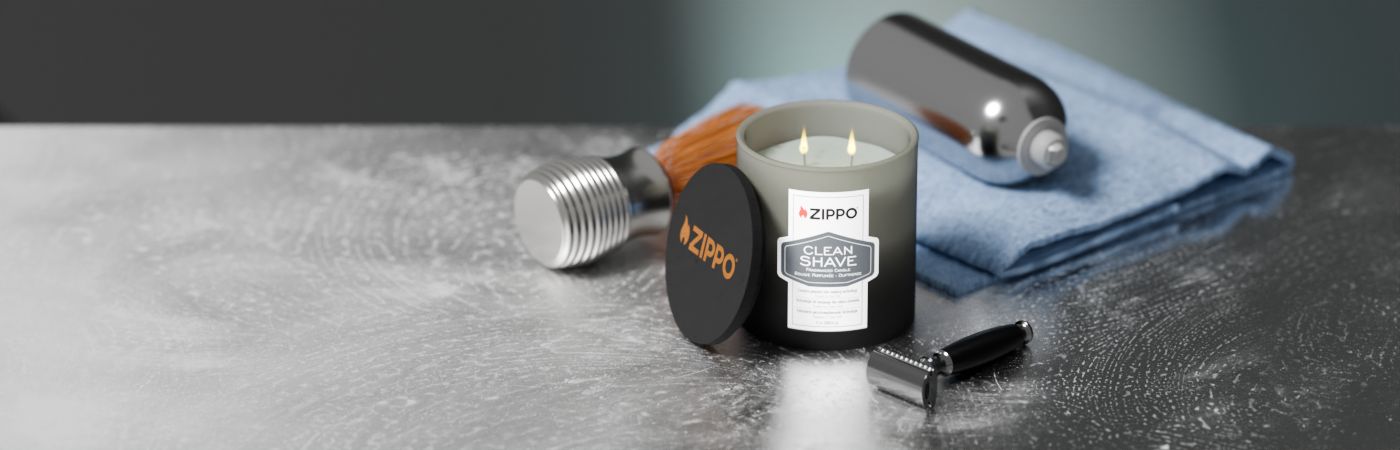Eine Zippo-Kerze mit der Aufschrift "Clean Shave" brennt mit zwei Flammen auf einem Tisch neben einem Rasierapparat, Handtuch und Accessoires auf einer grauen, strukturierten Oberfläche.